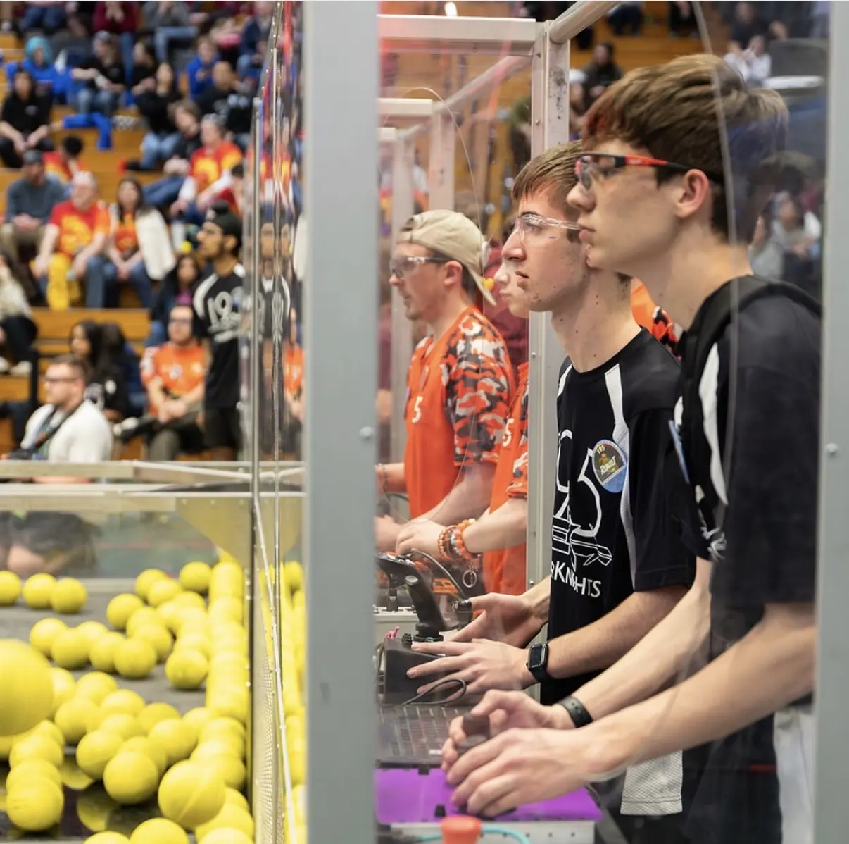 The Team 195 drive team at the controls behind the glass during a match, with the crowd in the stands behind them.