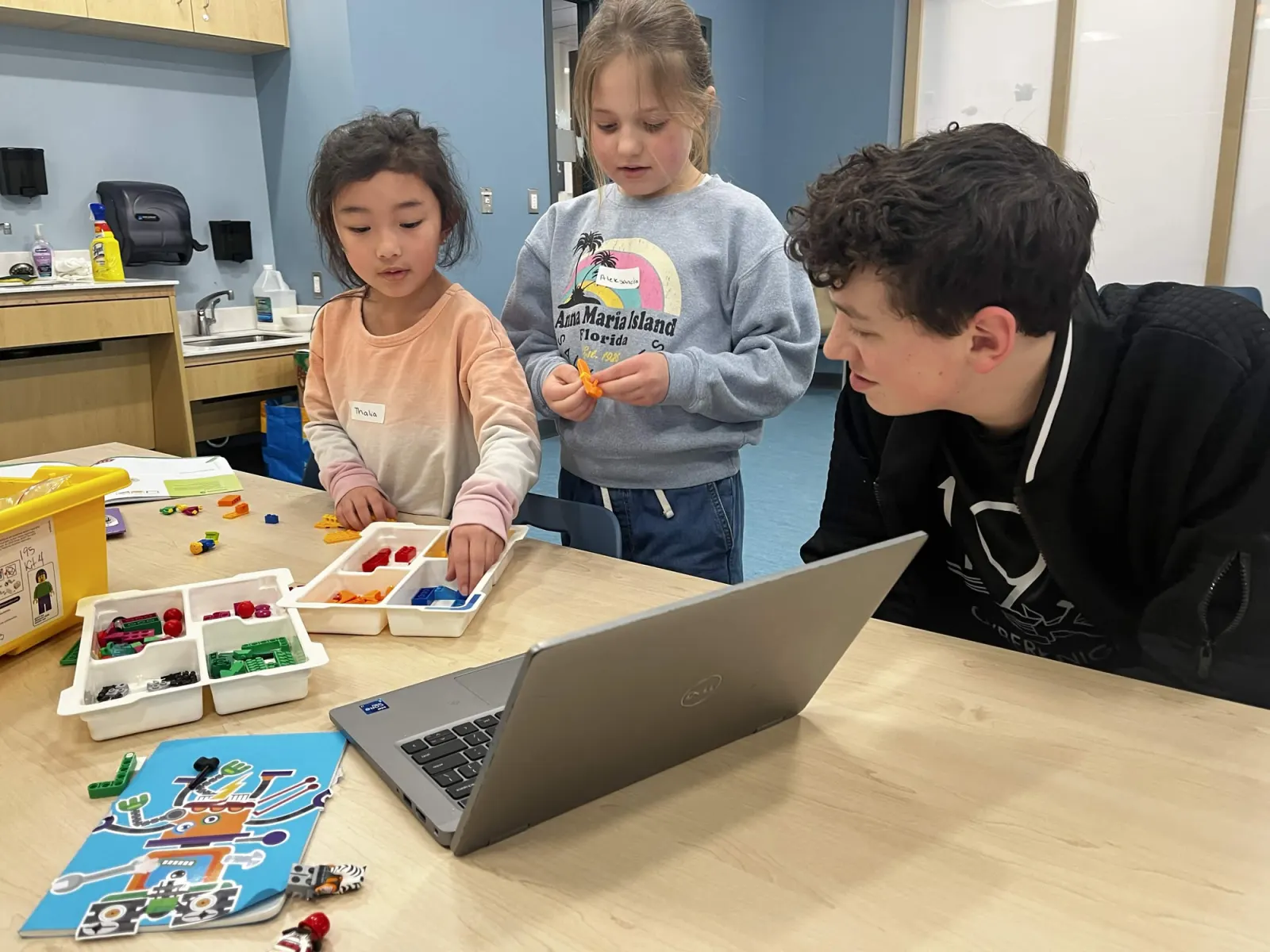 A Team 195 student coaching two young FIRST LEGO League students as they build with LEGO bricks beside a laptop.