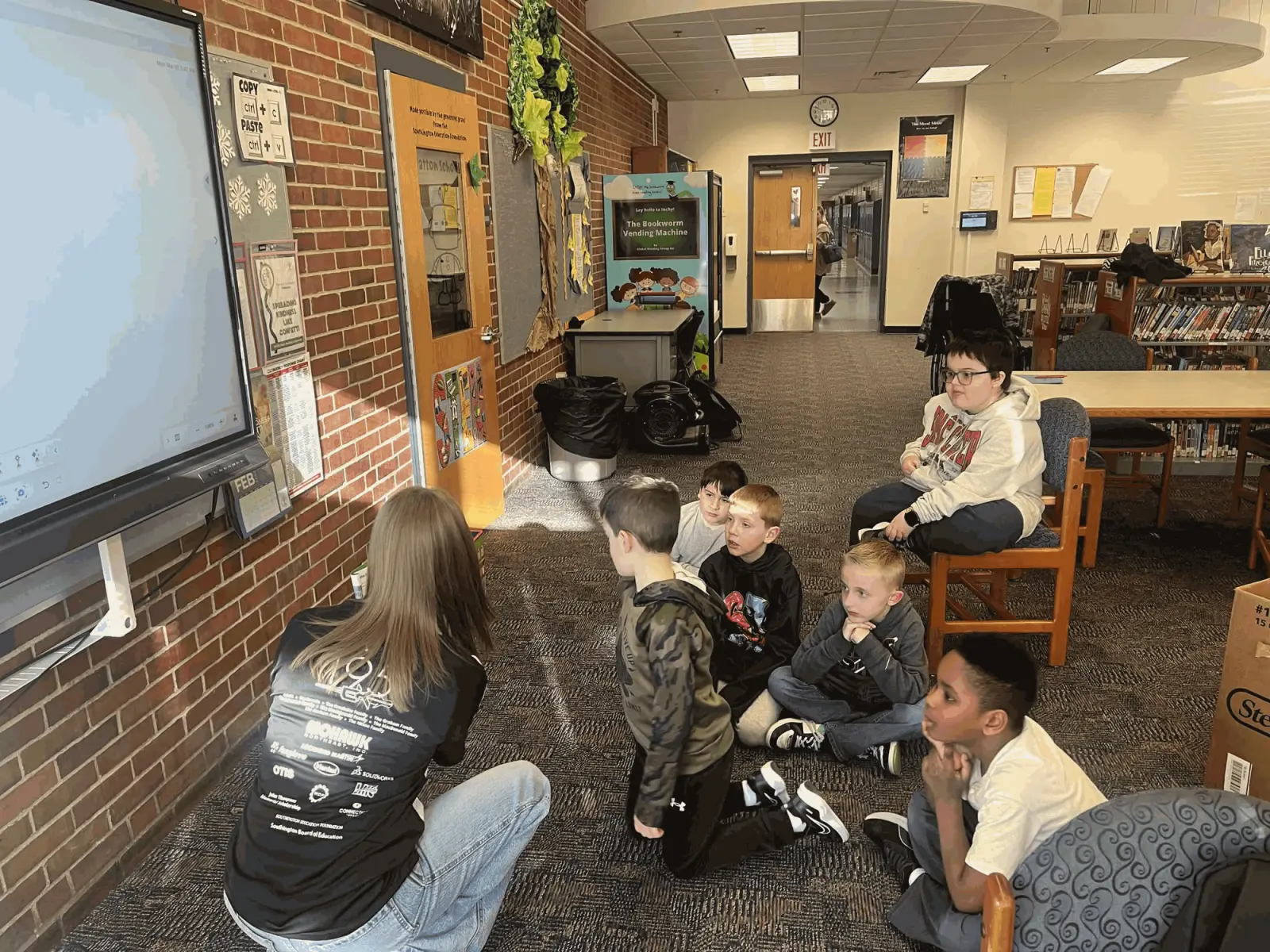 A Team 195 student coach leading a group of elementary school students in a school library.