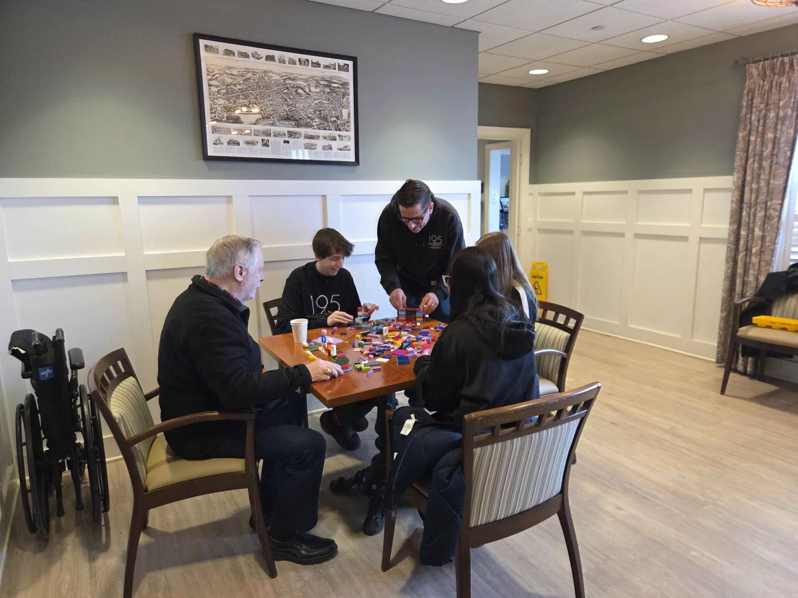 Team 195 students and a mentor building with LEGO bricks alongside a senior at an ElderKnights session.
