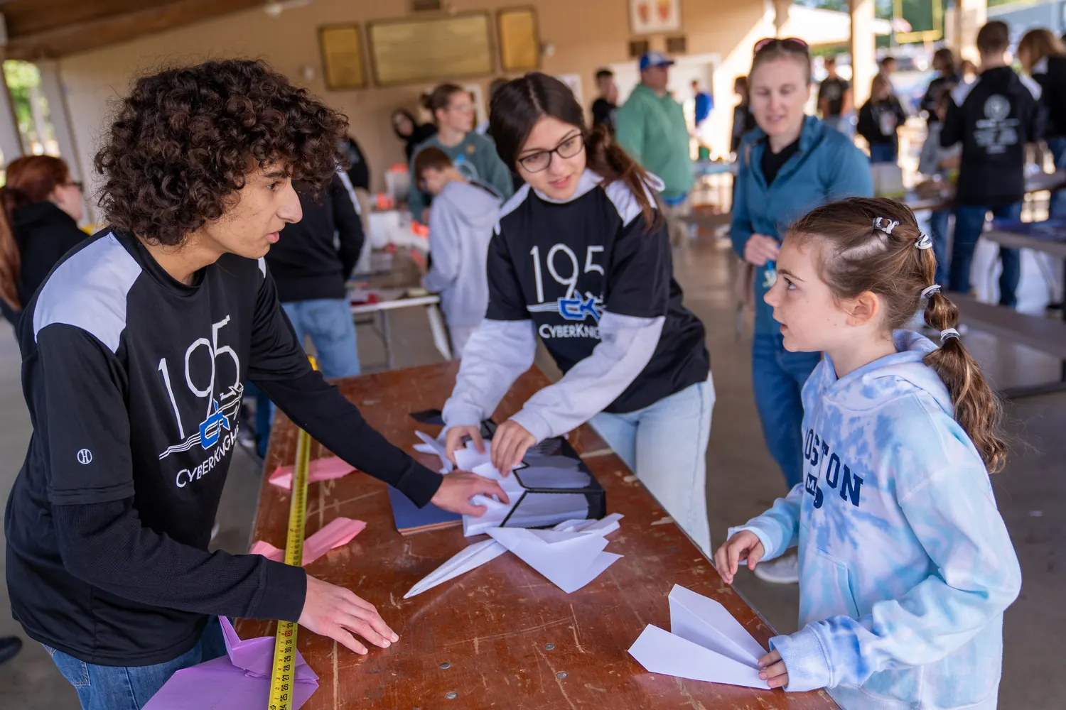 Team 195 students running a paper airplane station with children at an outdoor STEM event.