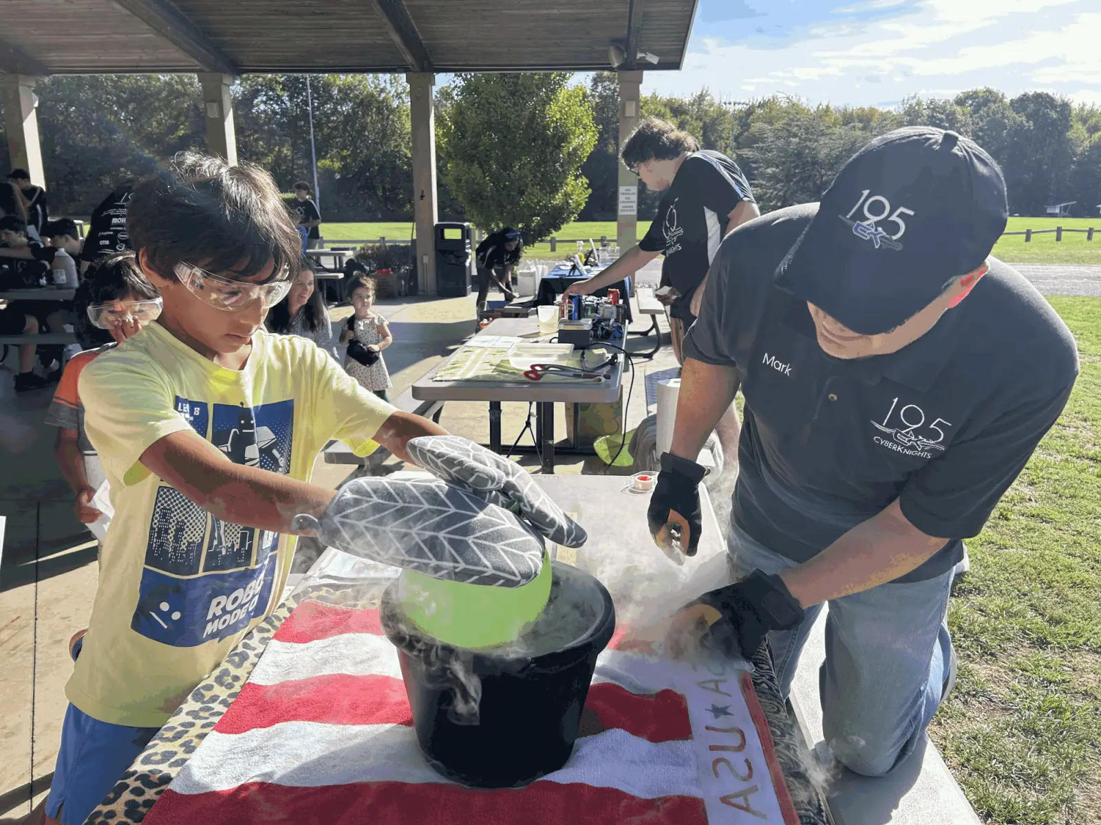 A child in safety glasses and oven mitts at a dry ice station at STEM For All, helped by a Team 195 mentor.
